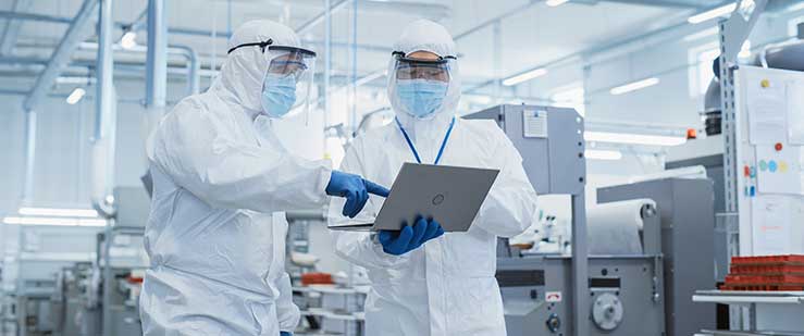 Two Scientists Walking in a Heavy Industry Factory in Sterile Coveralls and Face Masks, Using Laptop Computer