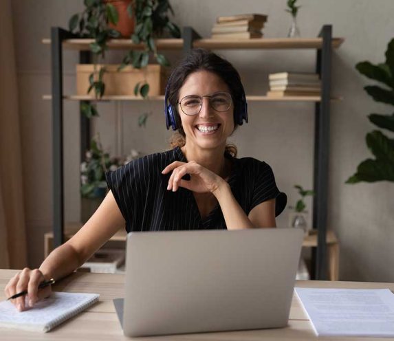 Head shot portrait of smiling woman in headphones using laptop, looking at camera, businesswoman studying online